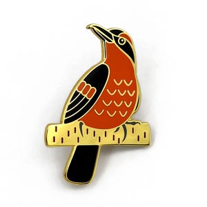 Könnte beinhalten: Goldfarbene Vogelbrosche mit rotem Körper, schwarzen Flügelakzenten und schwarzem Schwanz. Der Vogel sitzt auf einem goldfarbenen Ast. Die Brosche hat ein detailliertes Design und ist vor weißem Hintergrund platziert.