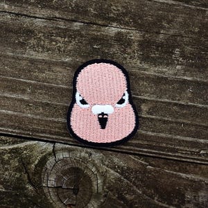 Puede incluir: Un parche bordado rosa con la imagen de una cara de pájaro estilizada con un contorno negro. El pájaro tiene un pico blanco y ojos enojados. El parche está sobre una superficie de madera envejecida.