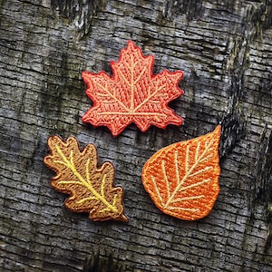 Może przedstawiać: Trzy haftowane naszywki w kształcie liści w jesiennych kolorach: pomarańczowym i żółtym. Naszywki, liść klonu, liść dębu i prosty kształt liścia, ułożone są na zwietrzałej, teksturowanej drewnianej powierzchni.