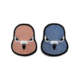 Puede incluir: Dos parches bordados con caras de pájaros. Uno es de color melocotón, el otro azul. Ambos tienen contornos negros, picos blancos y ojos enfadados. Estos parches son adecuados para ropa o accesorios.