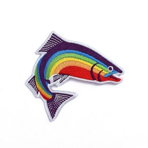 Puede incluir: Un parche bordado de una trucha arcoíris con un patrón de arcoíris en su espalda. La trucha es morada y roja con un contorno blanco.
