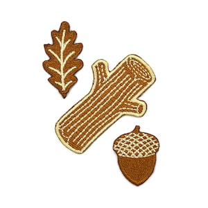 May include: Three embroidered patches featuring a brown oak leaf with cream stitching, a brown log with cream-colored bark texture lines, and a brown acorn with a textured cream cap. Each patch has a distinct shape and detailed thread work.
