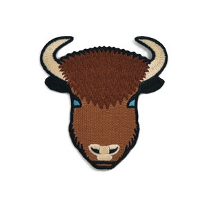 Op de afbeelding: Gestikte patch van een bruin bizonshoofd met blauwe ogen. De bizon kijkt naar voren met zijn hoorns omhoog.