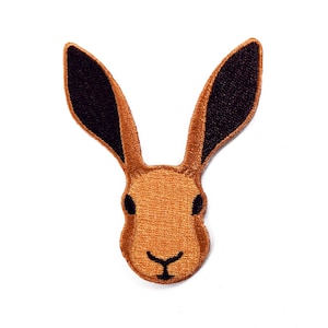 Könnte beinhalten: Gestickter Aufnäher mit einem Hasenkopf. Der Aufnäher hat einen braunen Körper mit schwarz gefärbten Ohrenspitzen und schwarzen Augen. Das Gesicht des Hasen ist mit einer kleinen, gebogenen schwarzen Linie für den Mund versehen.