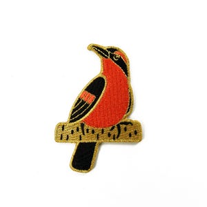 Könnte beinhalten: Gestickter Aufnäher mit einem stilisierten Vogel auf einem Ast. Der Vogel hat rotes Gefieder mit schwarzen Akzenten und einem schwarzen Schnabel. Der Ast ist goldfarben und strukturiert. Für Kleidung oder Accessoires.
