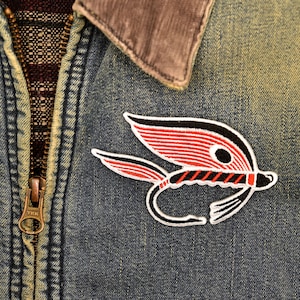 May include: A red and black embroidered patch of a fly fishing lure on a blue denim jacket.