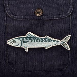 Könnte beinhalten: Ein blauer und weißer gestickter Aufnäher mit einem Makrelenfisch auf einem dunkelblauen Stoffhintergrund.