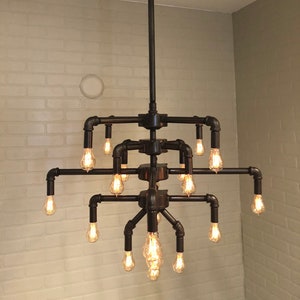 Puede incluir: Una lámpara de araña de tubería de metal negro con 12 bombillas Edison. La lámpara de araña tiene un diseño de varios niveles con múltiples brazos que se extienden desde un tubo central.