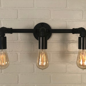 May include: Black industrial style wall sconce with three light bulbs. The sconce is made of black metal pipes and has a rustic look.