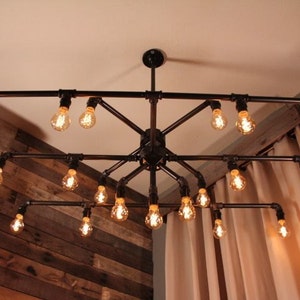 May include: A black metal pipe chandelier with 16 light bulbs. The chandelier is hanging from the ceiling and is surrounded by beige curtains.