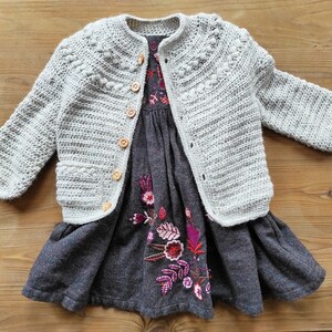May include: A gray crocheted cardigan with wooden buttons, worn over a gray dress with embroidered floral details in shades of pink and purple.