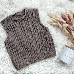 May include: A brown crocheted sleeveless vest with a ribbed texture. The vest is laid flat on a white fluffy surface.