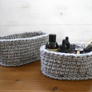 Może przedstawiać: Dwa szare owalne kosze na szydełku, jeden większy od drugiego. Mniejszy kosz zawiera kilka małych butelek i tubkę tuszu do rzęs z etykietą "Lashes".