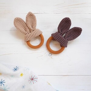 Puede incluir: Dos mordedores de madera para bebés con orejas de conejo de crochet. Un mordedor tiene orejas beige y el otro tiene orejas moradas.