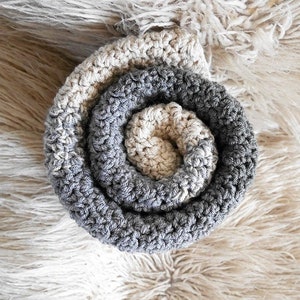 May include: A rolled, crocheted item in a spiral pattern. The item transitions from a light beige center to a medium gray outer edge. The texture is visible, with each stitch creating a raised, bumpy surface. The background is a soft, fluffy, off-white material.