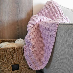 May include: A soft, purple crocheted throw blanket draped over a gray couch. The blanket has a textured, puffy design.