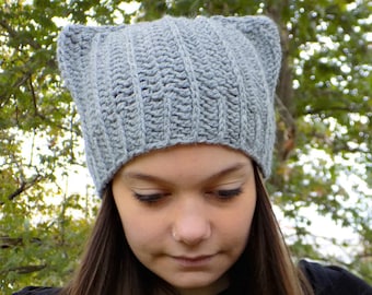 May include: A gray crocheted cat ear beanie with a ribbed texture. The beanie is worn by a person with long brown hair.