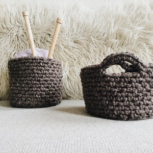 May include: Two brown crocheted baskets, one with a handle, on a white fuzzy surface. The smaller basket has a ball of purple yarn and two wooden knitting needles inside.