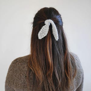 Puede incluir: Una mujer con cabello casta&ntilde;o lleva un accesorio para el pelo tejido de color gris claro con forma de orejas de conejo. Lleva un su&eacute;ter marr&oacute;n. El fondo es liso.