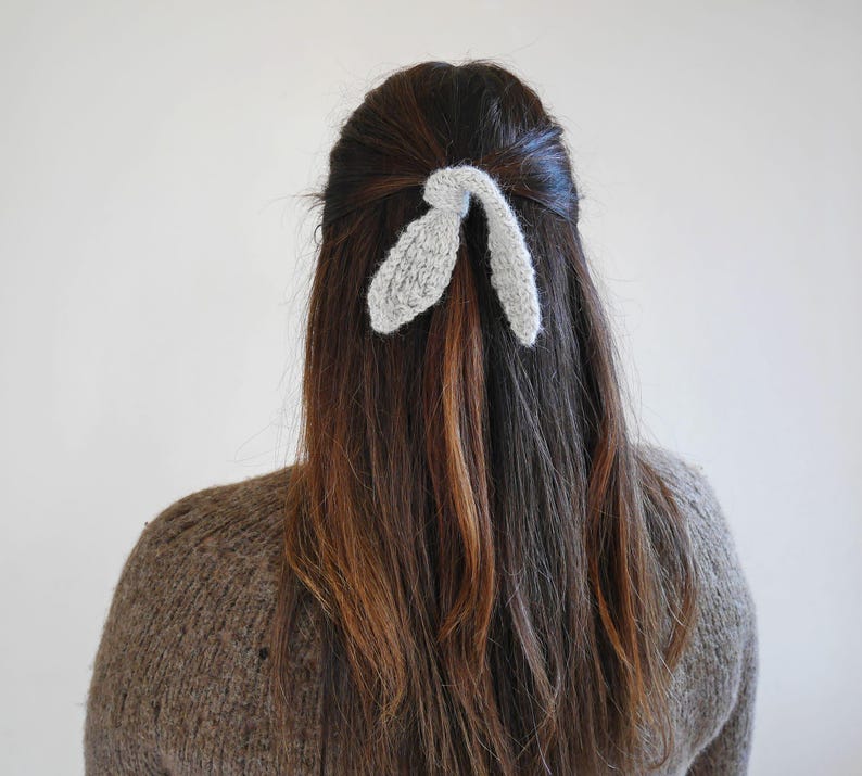 Puede incluir: Una mujer con cabello casta&ntilde;o lleva un accesorio para el pelo tejido de color gris claro con forma de orejas de conejo. Lleva un su&eacute;ter marr&oacute;n. El fondo es liso.