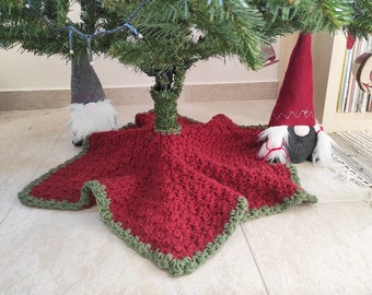 Puede incluir: Una falda de árbol de Navidad de crochet rojo con un borde verde. Dos gnomos están colocados en el suelo junto a la falda del árbol.