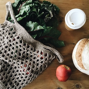 May include: A brown crocheted mesh bag filled with green leafy vegetables. A red apple, a loaf of bread, and a white takeaway coffee cup are on a wooden surface.