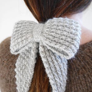Puede incluir: Un accesorio para el cabello en forma de lazo, tejido en gris claro. El lazo est&aacute; atado alrededor de una cola de caballo, con los extremos del lazo cayendo. El accesorio est&aacute; hecho de un hilo suave y texturizado.
