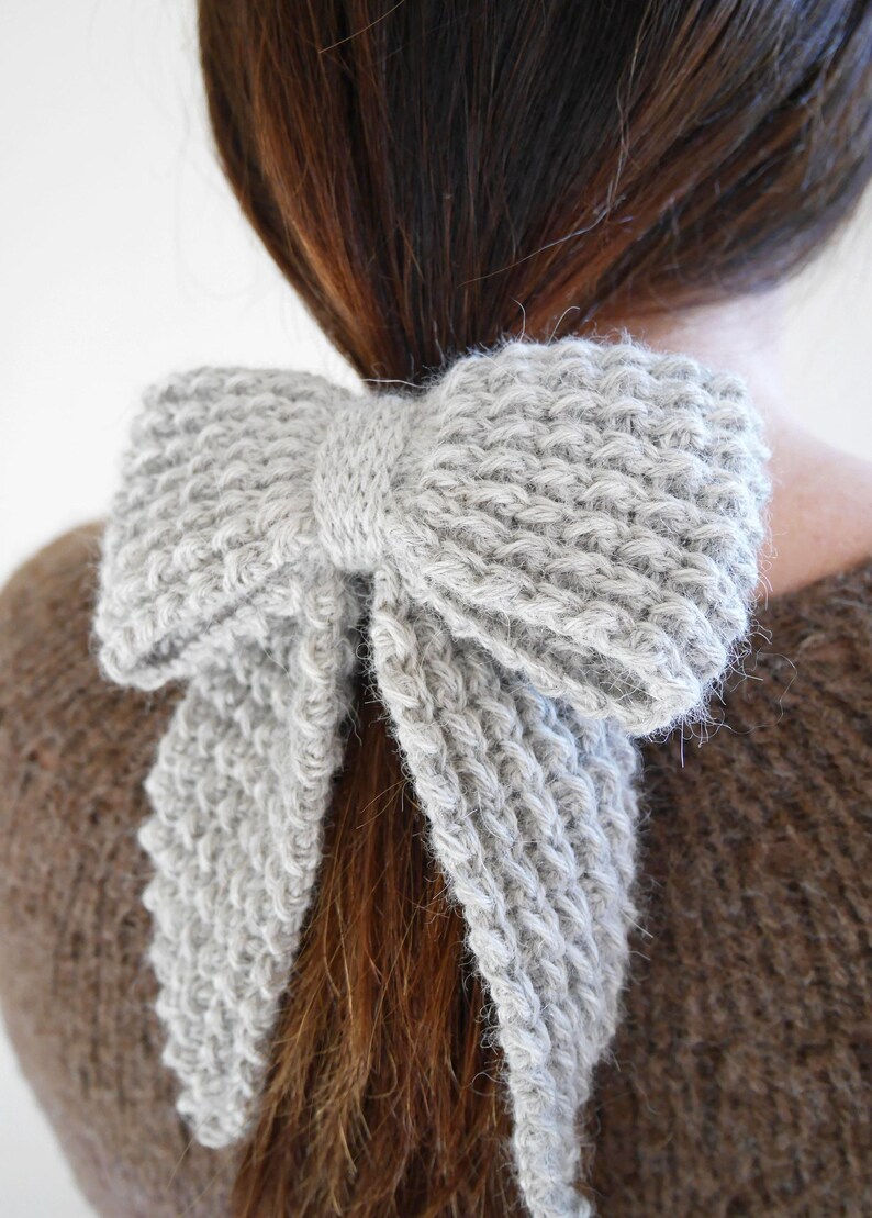 Puede incluir: Un accesorio para el cabello en forma de lazo, tejido en gris claro. El lazo est&aacute; atado alrededor de una cola de caballo, con los extremos del lazo cayendo. El accesorio est&aacute; hecho de un hilo suave y texturizado.