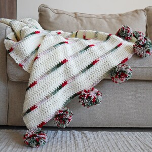 May include: A white crocheted throw blanket with red, green, and white pom-poms. The blanket is draped over a beige couch.