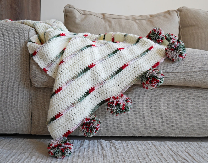 May include: A white crocheted throw blanket with red, green, and white pom-poms. The blanket is draped over a beige couch.