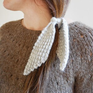 Puede incluir: Una goma para el pelo de punto gris claro est&aacute; envuelta alrededor de una coleta. La goma para el pelo tiene un aspecto texturizado y un borde festoneado decorativo. La persona lleva un su&eacute;ter marr&oacute;n con un punto texturizado.