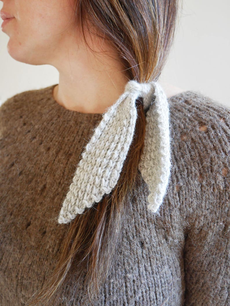 Puede incluir: Una goma para el pelo de punto gris claro est&aacute; envuelta alrededor de una coleta. La goma para el pelo tiene un aspecto texturizado y un borde festoneado decorativo. La persona lleva un su&eacute;ter marr&oacute;n con un punto texturizado.