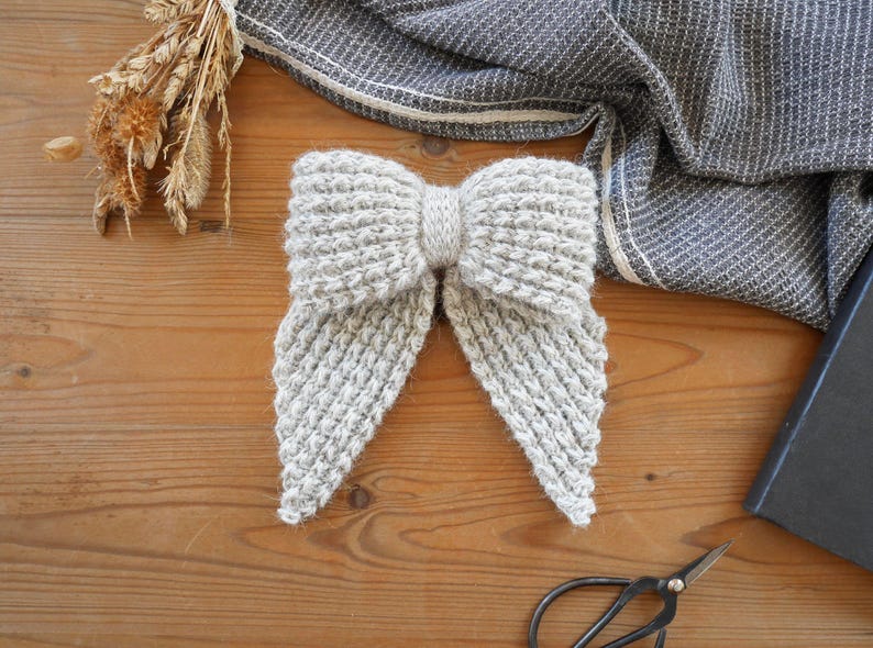 Puede incluir: Una pajarita de color gris claro tejida a mano con un patr&oacute;n texturizado. La pajarita est&aacute; hecha de hilo suave y tiene una forma de lazo cl&aacute;sica. La pajarita est&aacute; sobre una superficie de madera, con un pa&ntilde;o gris y unas tijeras en el fondo.