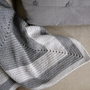 May include: A gray and white striped crocheted blanket with a textured pattern. The blanket is folded and draped over a light brown surface.