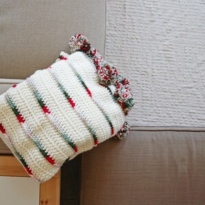 May include: A white crocheted pillow with red, green, and white stripes and pom-poms around the edges.