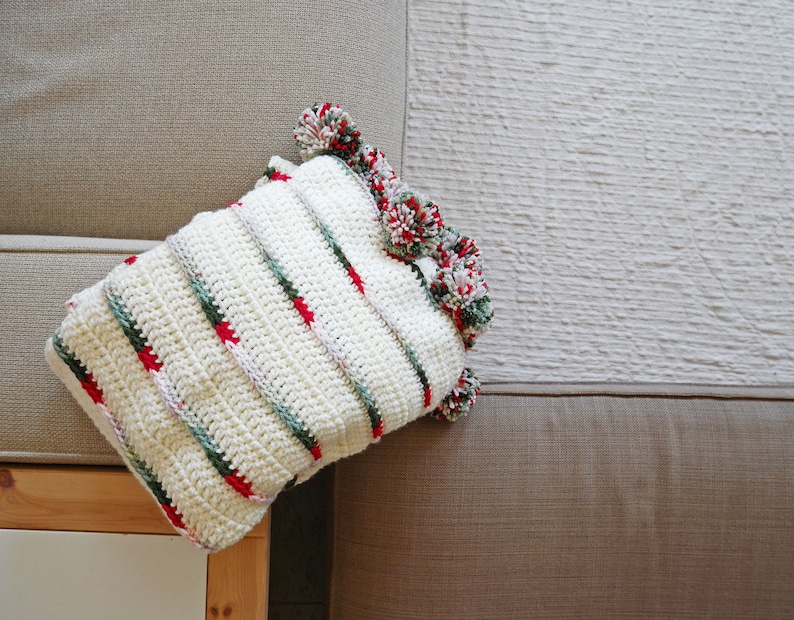 May include: A white crocheted pillow with red, green, and white stripes and pom-poms around the edges.