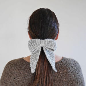 Puede incluir: Un accesorio para el cabello con forma de lazo tejido de color gris claro est&aacute; atado en una cola de caballo. El lazo est&aacute; hecho de una tela de punto texturizada y est&aacute; colocado en la parte posterior de la cabeza. La persona lleva un su&eacute;ter marr&oacute;n.