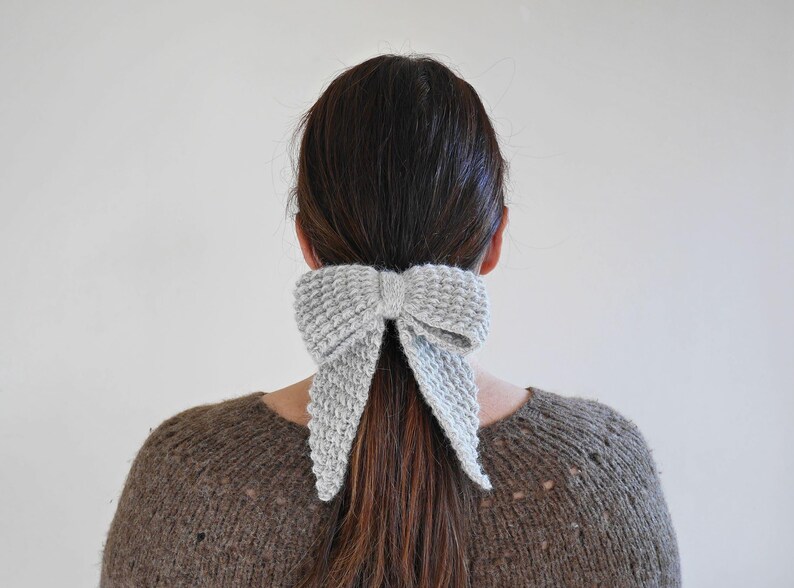 Puede incluir: Un accesorio para el cabello con forma de lazo tejido de color gris claro est&aacute; atado en una cola de caballo. El lazo est&aacute; hecho de una tela de punto texturizada y est&aacute; colocado en la parte posterior de la cabeza. La persona lleva un su&eacute;ter marr&oacute;n.