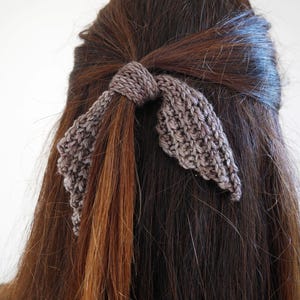 Puede incluir: Un accesorio para el cabello tejido en forma de lazo, en color gris neutro, est&aacute; atado en cabello casta&ntilde;o. El lazo est&aacute; hecho de una tela tejida texturizada, con los extremos del lazo colgando. El cabello es largo y liso, con una ligera onda.