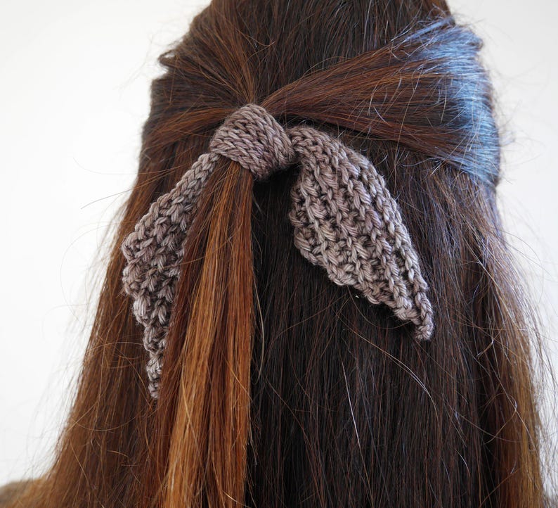 Puede incluir: Un accesorio para el cabello tejido en forma de lazo, en color gris neutro, est&aacute; atado en cabello casta&ntilde;o. El lazo est&aacute; hecho de una tela tejida texturizada, con los extremos del lazo colgando. El cabello es largo y liso, con una ligera onda.
