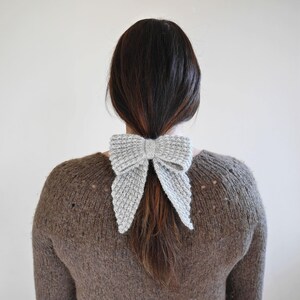 Puede incluir: Un accesorio para el cabello en forma de lazo, tejido en gris claro. El lazo est&aacute; atado alrededor de una cola de caballo. La persona lleva un su&eacute;ter marr&oacute;n. El fondo es una pared lisa de color blanco roto.