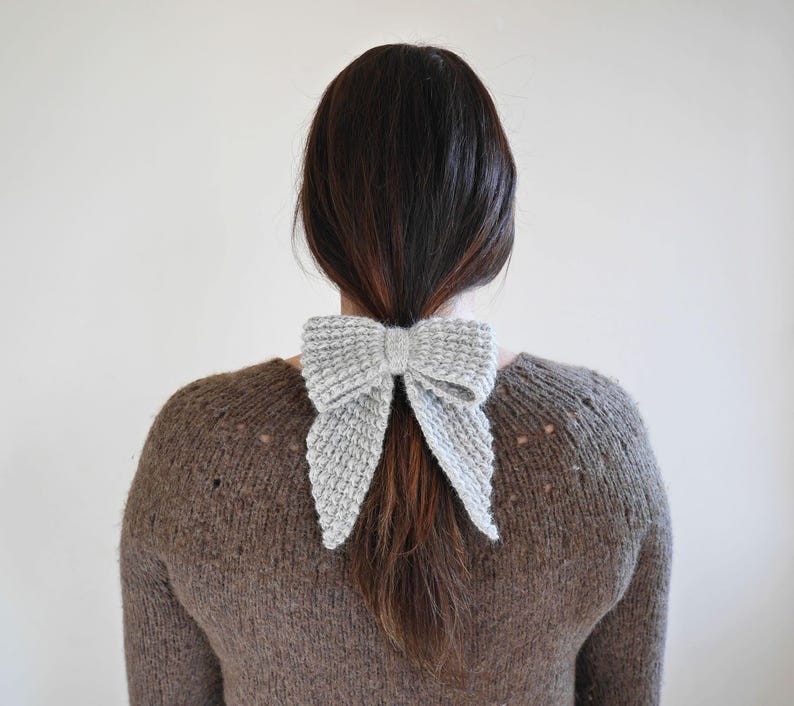 Puede incluir: Un accesorio para el cabello en forma de lazo, tejido en gris claro. El lazo est&aacute; atado alrededor de una cola de caballo. La persona lleva un su&eacute;ter marr&oacute;n. El fondo es una pared lisa de color blanco roto.