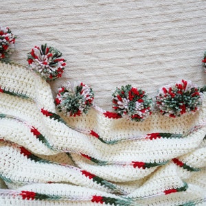 May include: A white crocheted blanket with green and red stripes. The blanket is decorated with pom-poms in shades of green, white, and red.