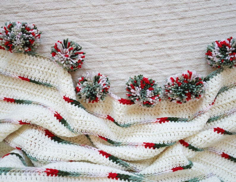 May include: A white crocheted blanket with green and red stripes. The blanket is decorated with pom-poms in shades of green, white, and red.