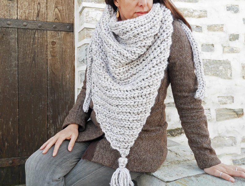 A woman with brown hair is sitting on a porch outside of a wooden door and wears jeans, a brown coat and an oversized crochet triangle shawl in grey color.