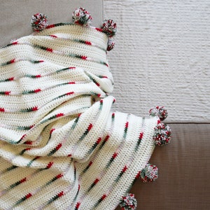 May include: A white crocheted throw blanket with red and green stripes and pom-poms on the edges.