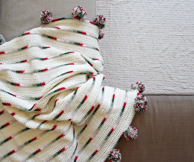 May include: A white crocheted throw blanket with red and green stripes and pom-poms on the edges.