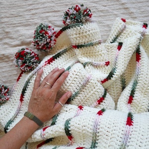 May include: White crochet blanket with red, green, and pink stripes and pom-poms around the edges.