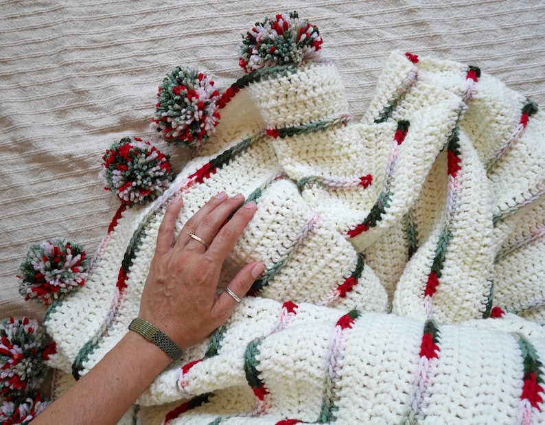 May include: White crochet blanket with red, green, and pink stripes and pom-poms around the edges.