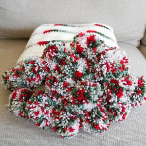May include: A white crocheted throw blanket with green and red pom-poms. The blanket is made with a striped pattern and has a soft, fluffy texture.